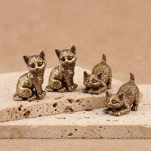 Può includere: Un set di quattro piccole statuette di gatto in ottone con texture del pelo dettagliate. Due gatti sono in posizione seduta eretta, mentre gli altri due sono in una posa accovacciata giocosa con le code sollevate verso l'alto.