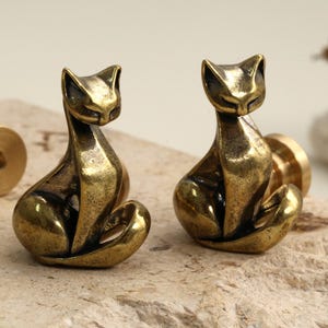 May include: Two brass cat figurines in a sitting pose. The cats have a stylized design with pointed ears and curved tails. The brass has a warm, aged patina. The figurines are on a light-colored, textured surface.