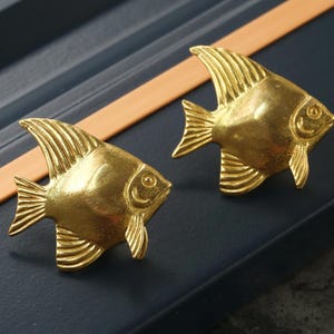 May include: Two gold-coloured fish-shaped drawer pulls. The fish are facing to the right and have detailed scales and fins.