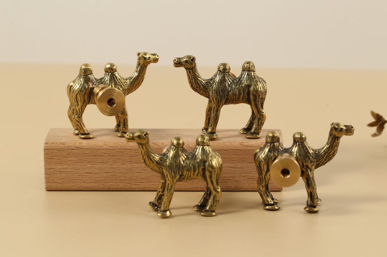 May include: Five antique brass camel figurines. The camels are standing on a wooden block. Each camel has a detailed design and a circular brass fitting on the back, suggesting they are drawer pulls or knobs.