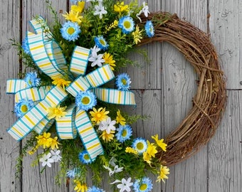 Op de afbeelding: Een lentekrans met blauwe en gele bloemen en een wit-blauwe gestreepte strik. De krans is gemaakt van druivenranken en hangt aan een grijze houten deur.