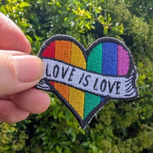 Op de afbeelding: Geborduurde patch in de vorm van een hart, met een regenboogontwerp en de witte tekst "LOVE IS LOVE". De patch heeft een zwarte rand en wordt getoond tegen een achtergrond van groen gebladerte.