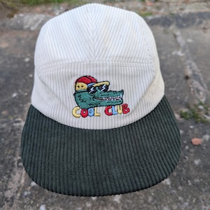 May include: Cream-colored corduroy cap with a dark green brim. The cap features an embroidered design of a green alligator wearing sunglasses and a red hat, with the words "COOL CLUB" in yellow and red.
