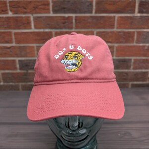 May include: A dusty rose baseball cap with a curved brim. The cap features the text "DOS & DOTS" above a yellow tiger graphic with white teeth. The hat is displayed on a glass head against a brick wall background.