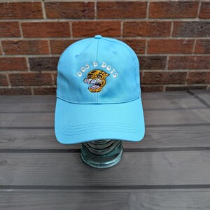 May include: A light blue baseball cap with white embroidered text reading "DOS & DOTS". The cap features a tiger graphic in yellow, orange, and black. The hat is made of a durable fabric and has a curved peak.