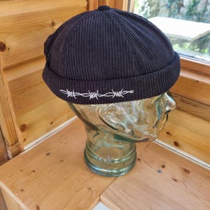 May include: A black corduroy beanie with a white barbed wire design embroidered around the bottom edge.