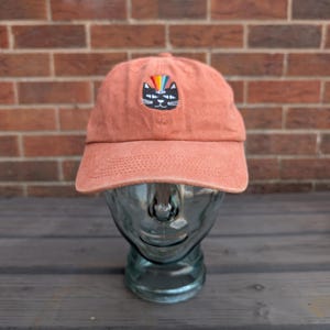 May include: A terracotta-coloured baseball cap featuring an embroidered graphic of a black cat face with a rainbow emanating from its forehead. The cap has a curved brim and a structured crown, displaying a simple, clean design on the front panel.