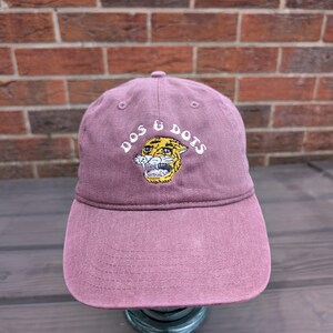 May include: A mauve baseball cap with the text "DOS & DOTS" embroidered above a tiger graphic. The cap has a curved brim and a fabric strap closure. The hat is made of a soft, durable material.