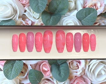 May include: A set of 10 pink and light pink press-on nails with a heart design. The nails are in a box with a white background.