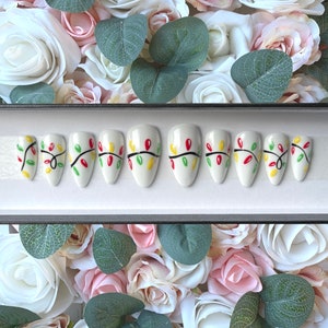 Peut inclure: Ensemble de dix faux ongles blancs avec un motif de lumières de Noël. Les ongles sont décorés de lumières vertes, rouges et jaunes.