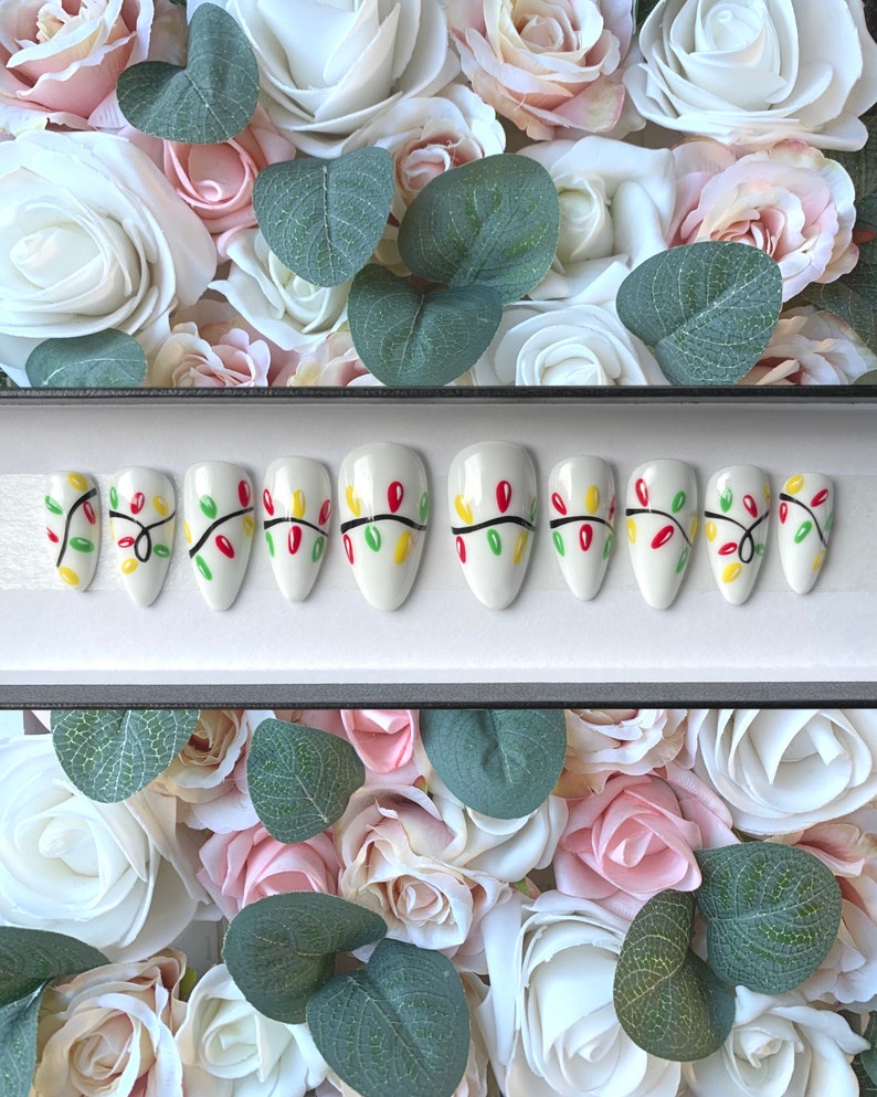 May include: Set of ten white press-on nails with a Christmas lights design. The nails are decorated with green, red, and yellow lights.