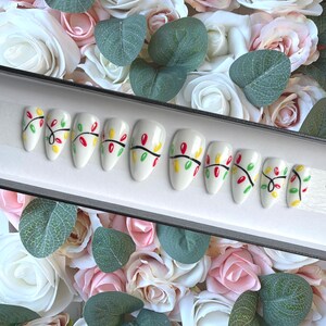 May include: A set of ten white false nails with a Christmas lights design. The nails are decorated with red, green, and yellow lights and black lines.