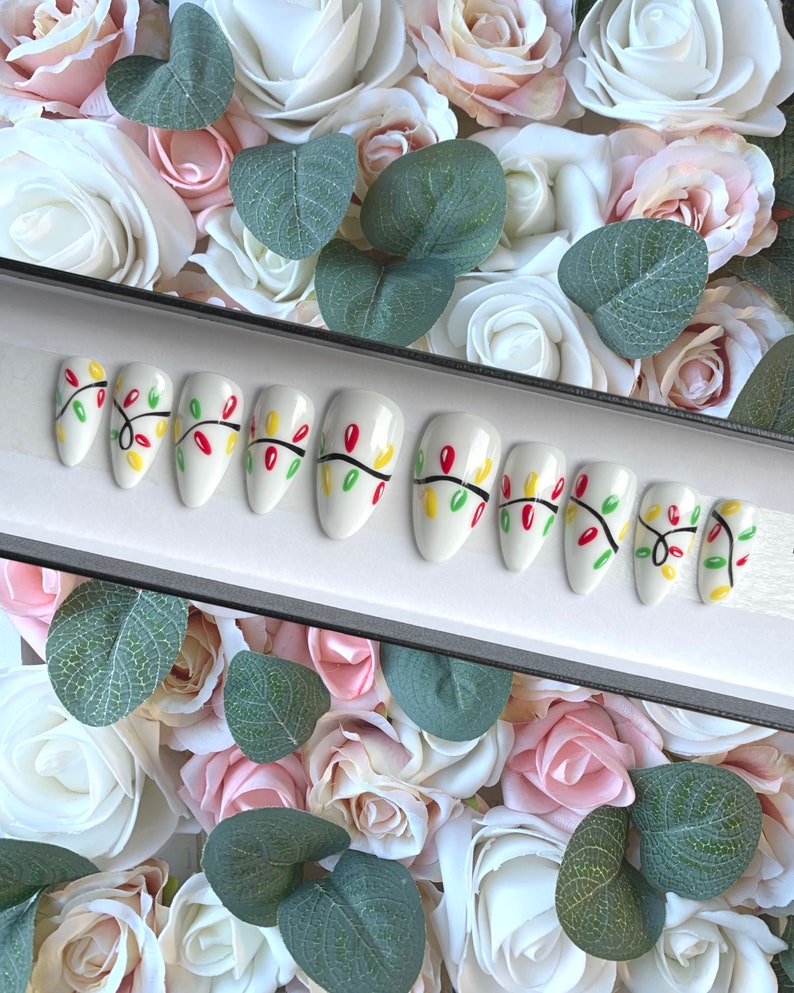 May include: A set of ten white false nails with a Christmas lights design. The nails are decorated with red, green, and yellow lights and black lines.