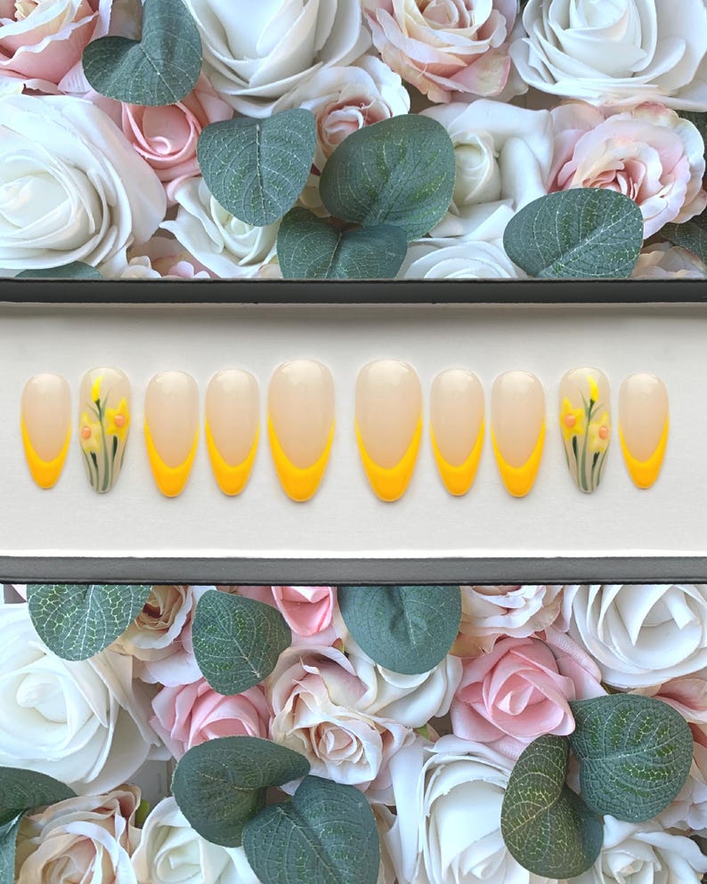 May include: A set of ten false nails with a clear base and a yellow tip. Each nail is decorated with a small, hand-painted daffodil.