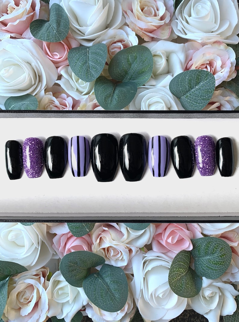 May include: A set of ten false nails in a black coffin shape. The nails are decorated with black and purple stripes, glitter, and solid black polish.