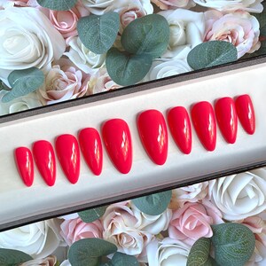May include: A set of ten bright red, almond-shaped, fake nails in a white box. The nails are arranged in two rows of five. The box is sitting on a bed of pink and white roses.