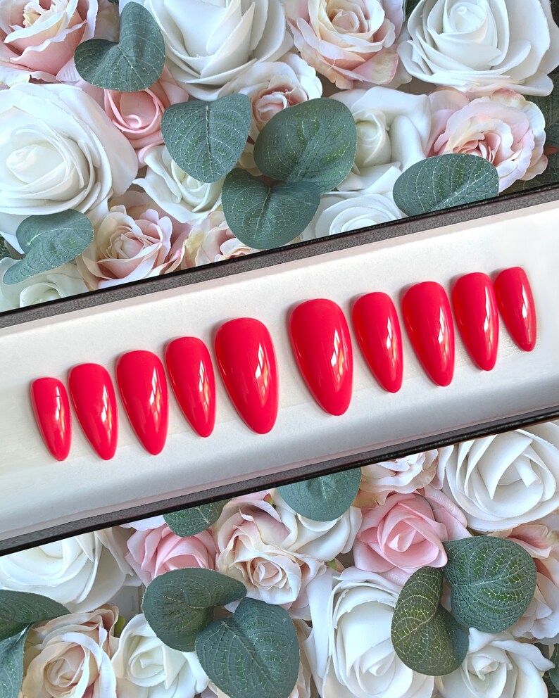 May include: A set of ten bright red, almond-shaped, fake nails in a white box. The nails are arranged in two rows of five. The box is sitting on a bed of pink and white roses.