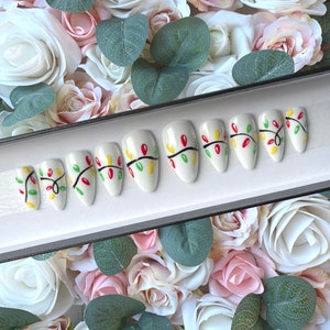 May include: A set of ten white false nails with a Christmas lights design. The nails are decorated with red, green, and yellow lights on a black wire.