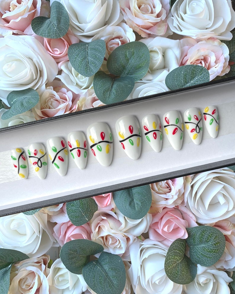May include: A set of ten white false nails with a Christmas lights design. The nails are decorated with red, green, and yellow lights on a black wire.