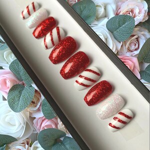 Puede incluir: Conjunto de diez u&ntilde;as postizas de tem&aacute;tica de bast&oacute;n de caramelo rojo y blanco. Las u&ntilde;as est&aacute;n decoradas con purpurina roja y rayas blancas.