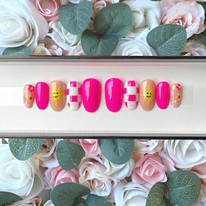 Puede incluir: Un conjunto de diez uñas postizas rosas con diferentes diseños. Los diseños incluyen un patrón a cuadros, un sol sonriente y un grupo de flores rosas.