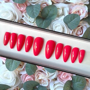 May include: A set of ten bright red almond-shaped fake nails in a white box. The nails are arranged in two rows of five. The box is surrounded by white and pink roses.