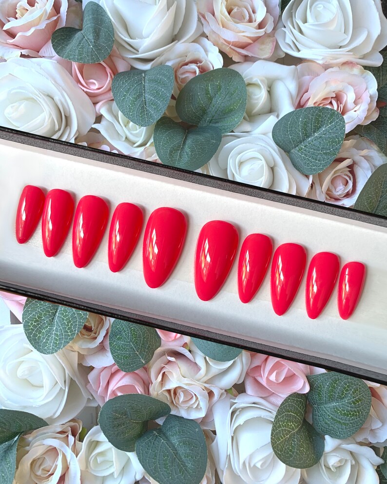 May include: A set of ten bright red almond-shaped fake nails in a white box. The nails are arranged in two rows of five. The box is surrounded by white and pink roses.
