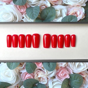 May include: A set of ten red coffin-shaped false nails in a black box. The nails are arranged in two rows of five. The box is sitting on a bed of white and pink roses.