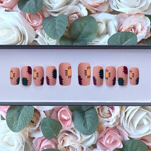 Könnte beinhalten: Ein Set aus zehn quadratischen Press-On-Nägeln. Jeder Nagel ist hautfarben mit einem Patchwork-Design in Dunkelgrün, Burgunderrot und Gelb-Orange. Der Hintergrund zeigt weiße und rosa Rosen und grüne Blätter.