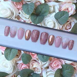 Pu&ograve; includere: Un set di dieci unghie finte in uno schema di colori rosa e oro. Le unghie sono disposte in una scatola con sfondo bianco. Alcune unghie hanno una finitura glitterata, mentre altre hanno una finitura metallica.