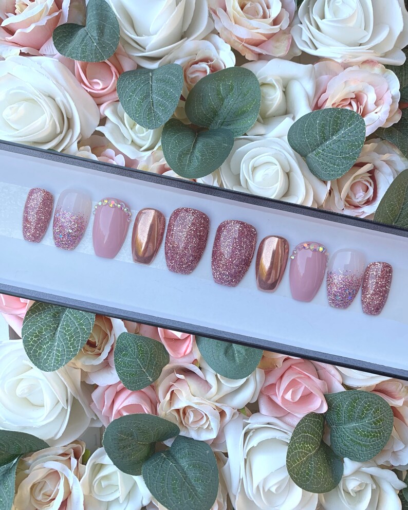 Pu&ograve; includere: Un set di dieci unghie finte in uno schema di colori rosa e oro. Le unghie sono disposte in una scatola con sfondo bianco. Alcune unghie hanno una finitura glitterata, mentre altre hanno una finitura metallica.