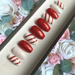 Puede incluir: Conjunto de diez u&ntilde;as postizas de purpurina roja y blanca con un dise&ntilde;o de bast&oacute;n de caramelo. Las u&ntilde;as se muestran en una bandeja negra con fondo blanco.
