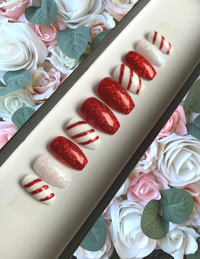 Puede incluir: Conjunto de diez u&ntilde;as postizas de purpurina roja y blanca con un dise&ntilde;o de bast&oacute;n de caramelo. Las u&ntilde;as se muestran en una bandeja negra con fondo blanco.