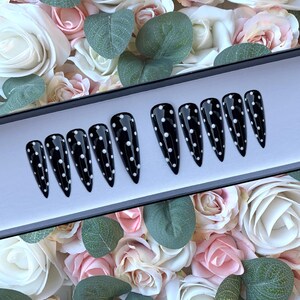 May include: A set of ten black stiletto false nails with white polka dots. The nails are displayed in a black box on a background of pink and white roses.