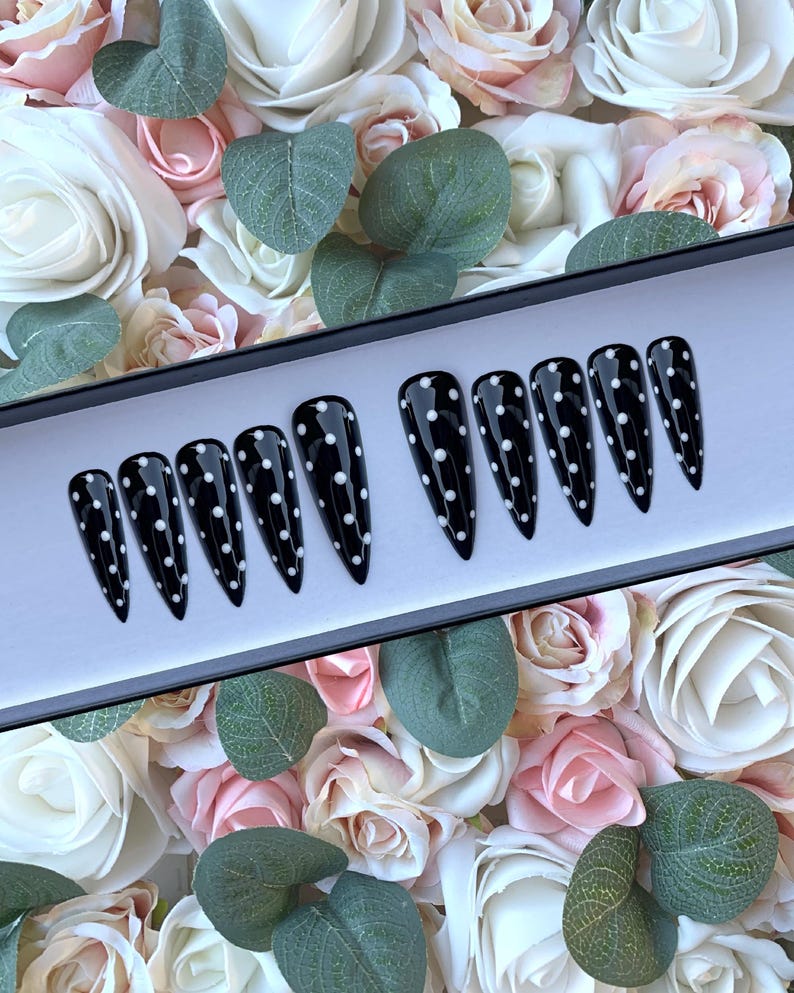May include: A set of ten black stiletto false nails with white polka dots. The nails are displayed in a black box on a background of pink and white roses.
