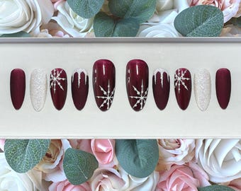 May include: A set of ten artificial nails in a variety of designs. The nails are a deep burgundy, white glitter, and burgundy with white snowflake designs. The nails are displayed against a backdrop of white and pink roses and green leaves.