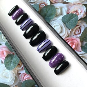 May include: A set of ten false nails in a black coffin shape. The nails are decorated with purple glitter, black and white stripes, and a solid black polish.