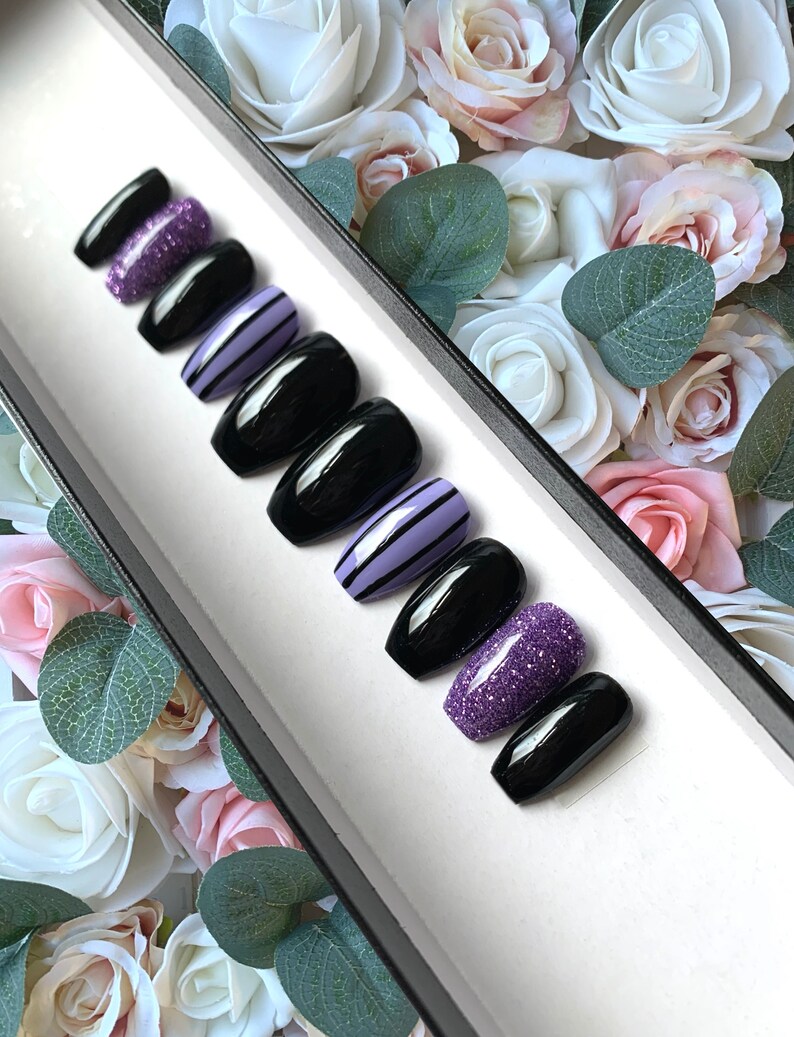 May include: A set of ten false nails in a black coffin shape. The nails are decorated with purple glitter, black and white stripes, and a solid black polish.
