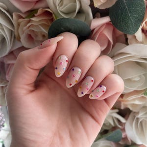 May include: A hand displaying long, almond-shaped nails. The nails are painted with a sheer, nude polish and decorated with colourful, sparkling rhinestones in a polka dot design. Soft pink and white roses form the background.