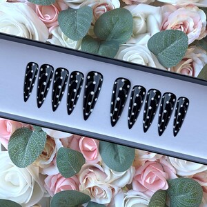 May include: A set of ten black stiletto false nails with white polka dots. The nails are displayed in a black box with a white interior.