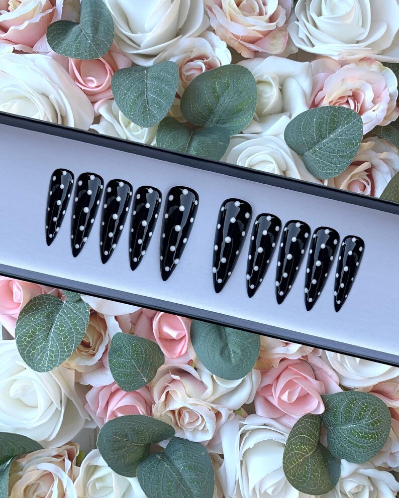 May include: A set of ten black stiletto false nails with white polka dots. The nails are displayed in a black box with a white interior.