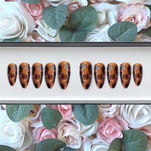 Puede incluir: Un conjunto de diez uñas postizas de color marrón y dorado con forma de ataúd.