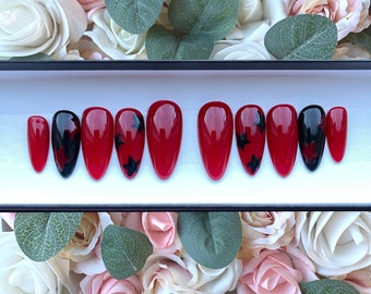 May include: A set of ten red and black false nails with a star design. The nails are arranged in a row in a black box.