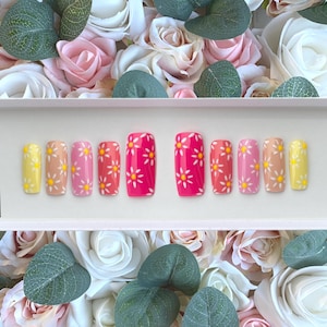 May include: A collection of press-on nails in shades of pink and yellow, decorated with white daisy patterns. The nails are displayed in a row, highlighting the different colours and designs. The background includes white, pink, and green floral arrangements.