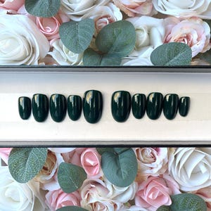 May include: A set of ten dark green oval-shaped press-on nails in a white box. The nails are arranged in two rows of five. The nails are surrounded by white and pink flowers.