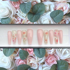 May include: A set of ten pink press-on nails with a floral design. The nails are in a white box with a black lid.