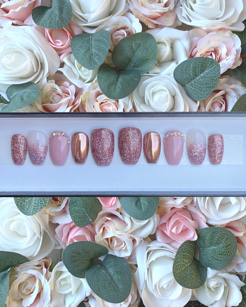 Pu&ograve; includere: Un set di dieci unghie finte con un designo di glitter rosa e oro. Le unghie sono disposte in una scatola con sfondo bianco.