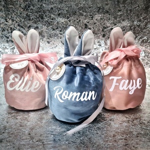 May include: Three velvet drawstring bags with bunny ears, each with a different name embroidered on the front: Ellie, Roman, and Faye.