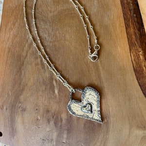 May include: A silver chain necklace with a large, textured silver heart pendant. The heart has a smaller heart cutout in the center.