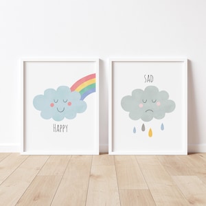 May include: Two framed art prints. One shows a smiling blue cloud with a rainbow and the word "HAPPY". The other depicts a sad grey cloud with raindrops and the word "SAD". Both are in white frames.
